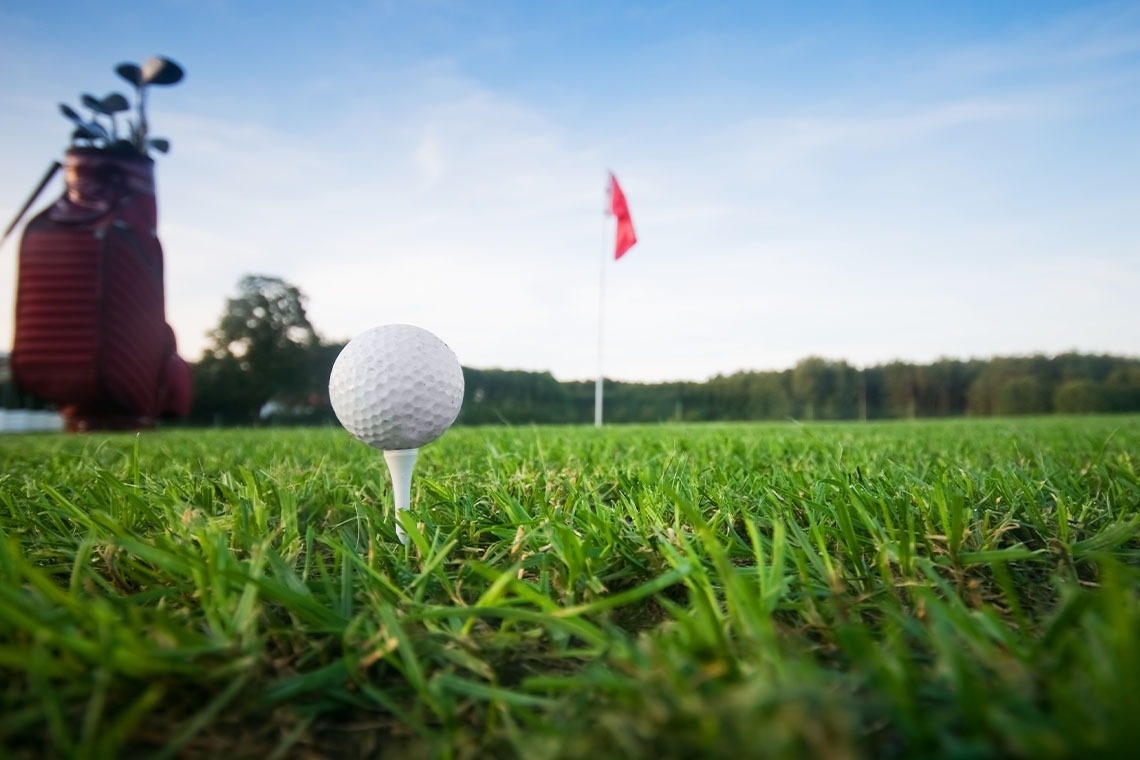 Ein Mann und eine Frau gehen mit ihren Golfausrüstungen über einen grünen Golfplatz, mit Bäumen im Hintergrund.