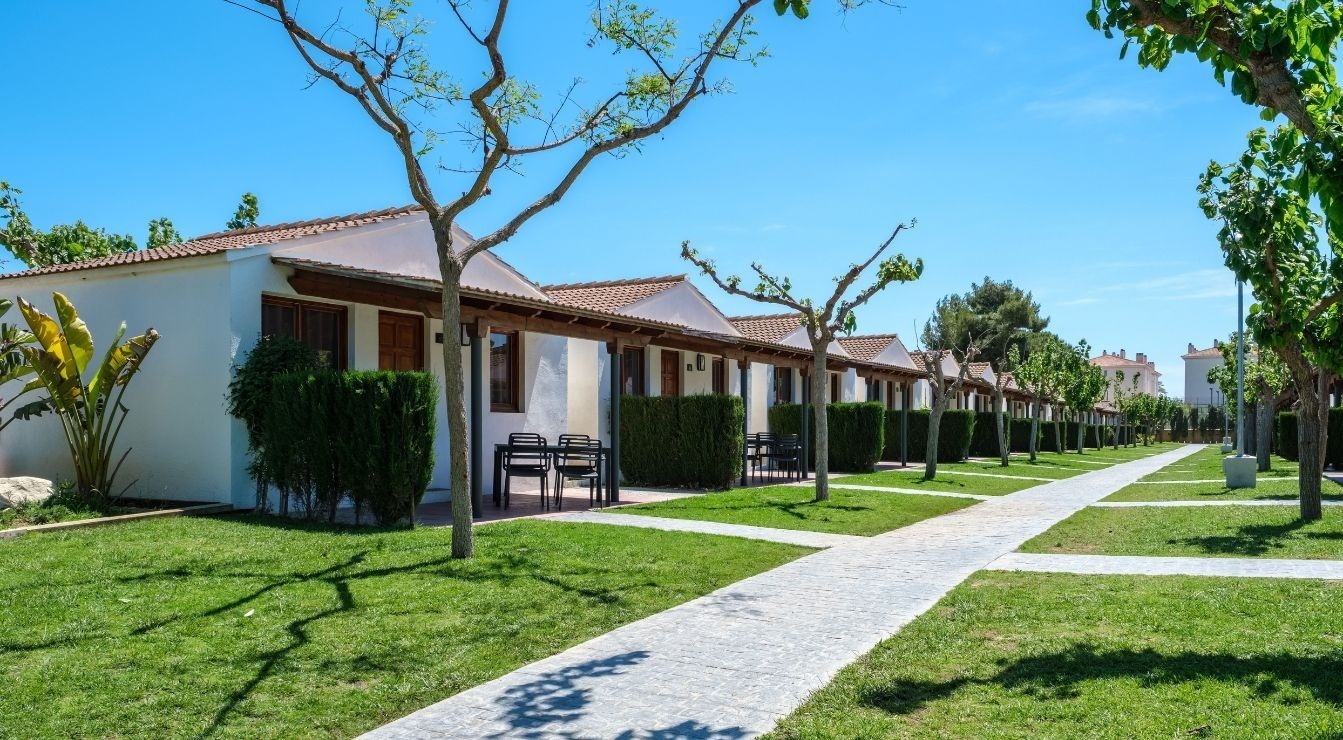 Una vista assolellada d'una filera de bungalous blancs amb teulades de teula, envoltats de gespa verda, arbres i camins pavimentats en un entorn de vacances.
