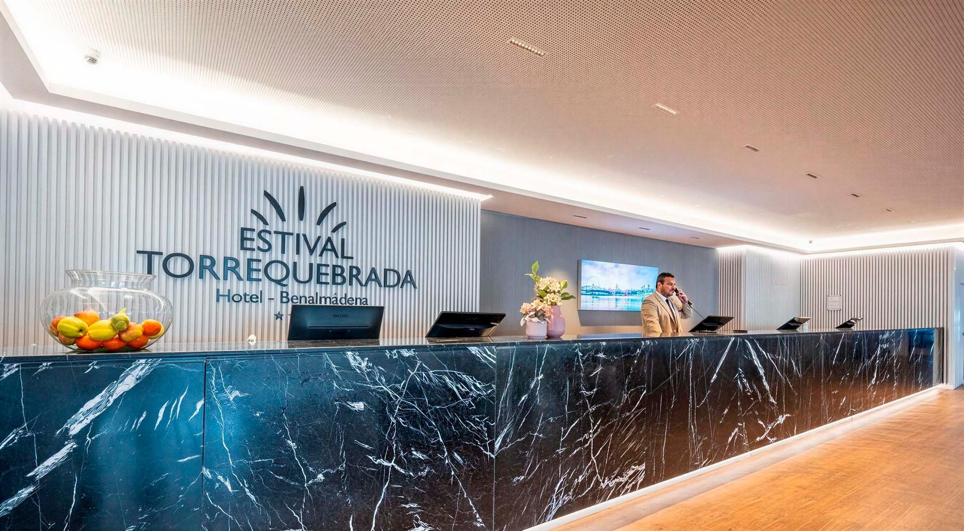 A modern hotel reception features a black marble front desk where a staff member is on the phone, with the "Estival Torrequebrada" logo prominently displayed on the textured wall behind him.