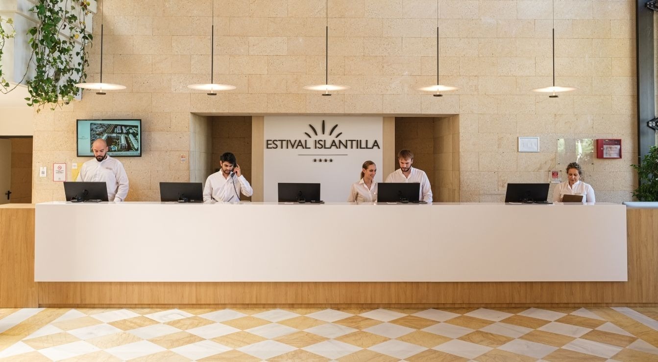 La imagen muestra la recepción de un hotel moderno con cinco recepcionistas trabajando detrás de un mostrador blanco, con el logo "ESTIVAL ISLANTILLA ****" de fondo.