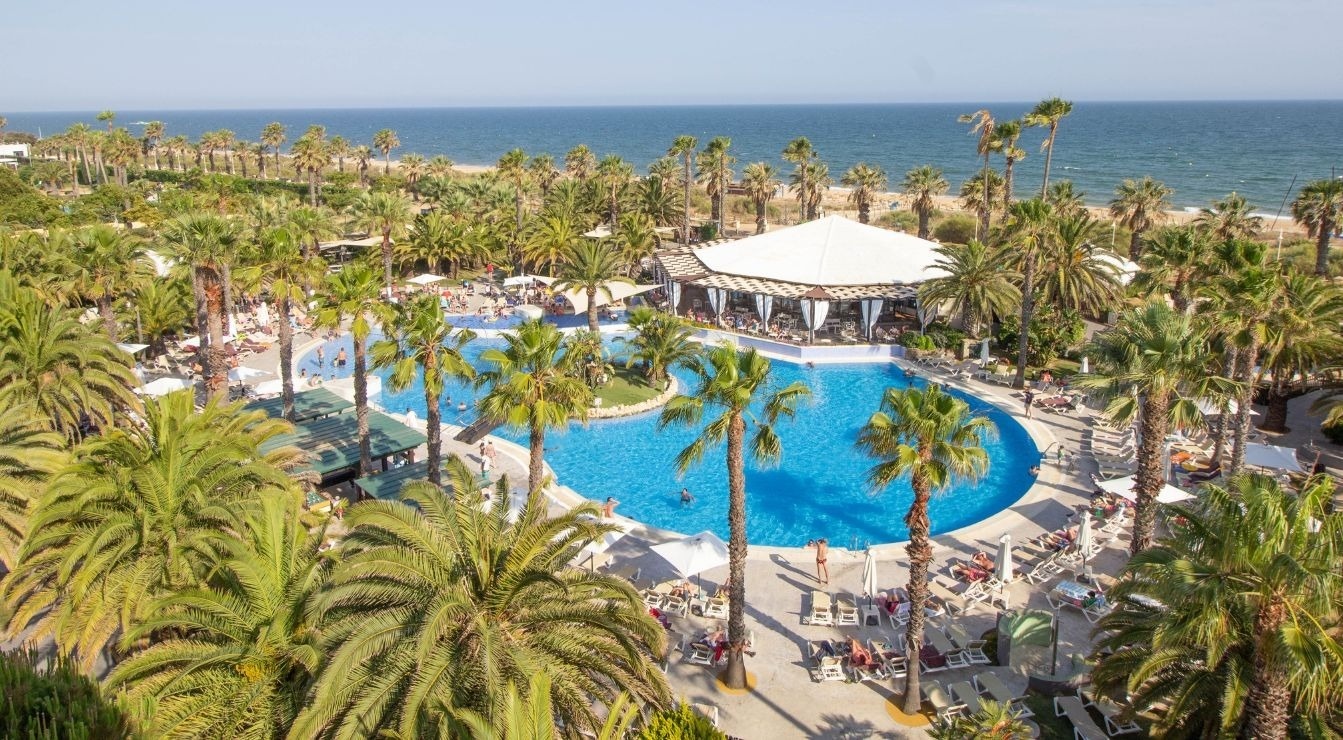An aerial view captures a vibrant resort with a large, blue swimming pool surrounded by numerous palm trees, a white-canopied restaurant, and a sandy beach leading to the clear ocean under a sunny sky.