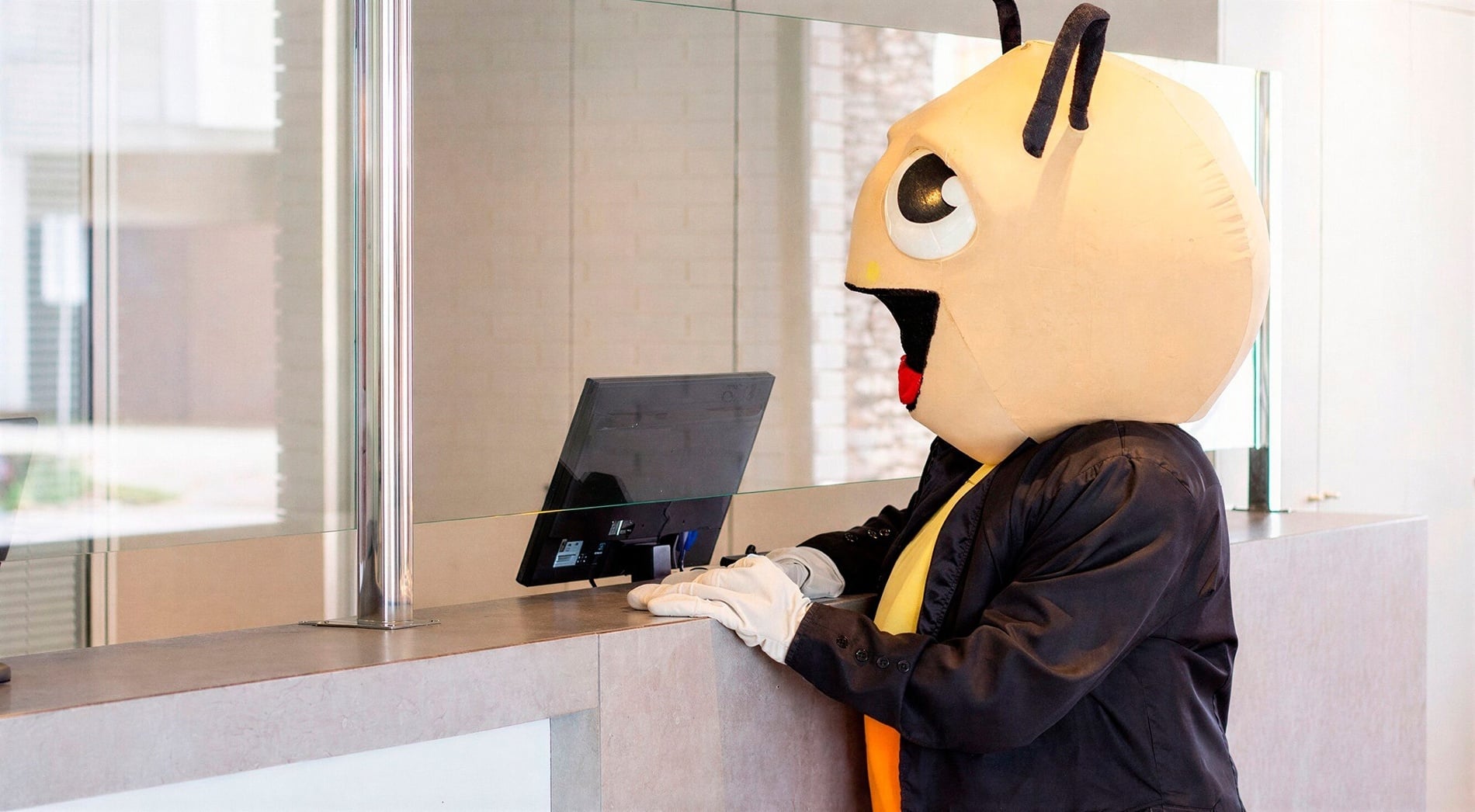 A person in an insect mascot costume, dressed in a suit and white gloves, stands at a counter and looks at a computer screen behind a glass partition.