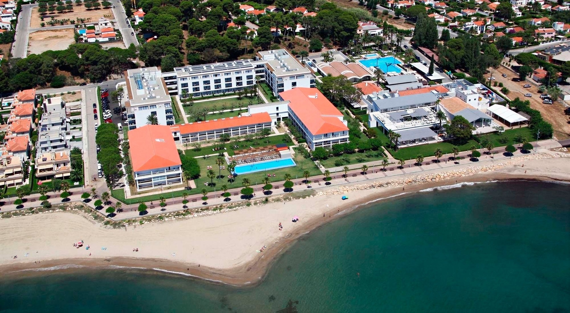 An aerial view captures a coastal resort complex featuring white buildings, orange roofs, multiple swimming pools, green lawns, a sandy beach, and clear blue sea.