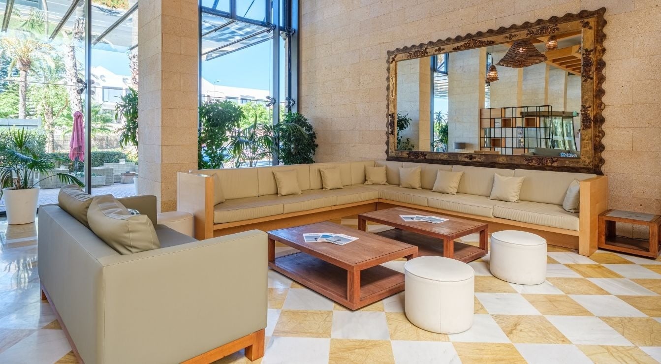 Un amplio y luminoso salón moderno, con sofás claros, mesas de madera, un gran espejo decorativo y suelo de baldosas a cuadros, ofrece vistas a un jardín a través de sus grandes ventanales.