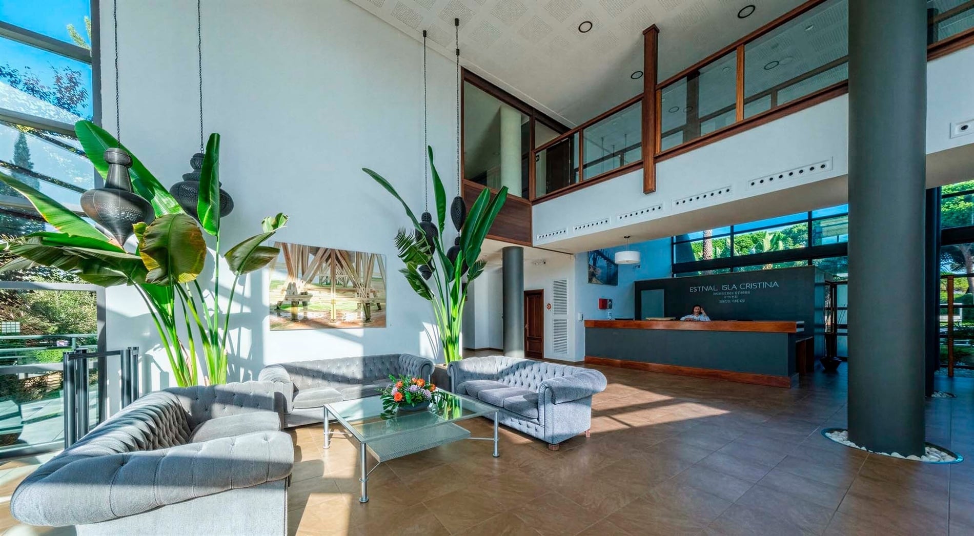 Un hall d'hôtel moderne et lumineux présente un coin salon confortable avec des canapés gris, de grandes plantes vertes et un comptoir de réception au fond.