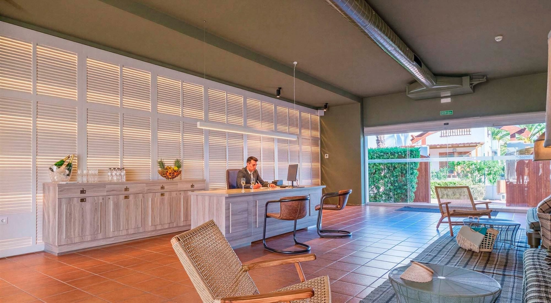 A modern reception area, possibly a hotel lobby, features a man working at a light-colored desk in front of an illuminated slatted wall, with terracotta tiled flooring and a glass door leading to an outdoor garden view.