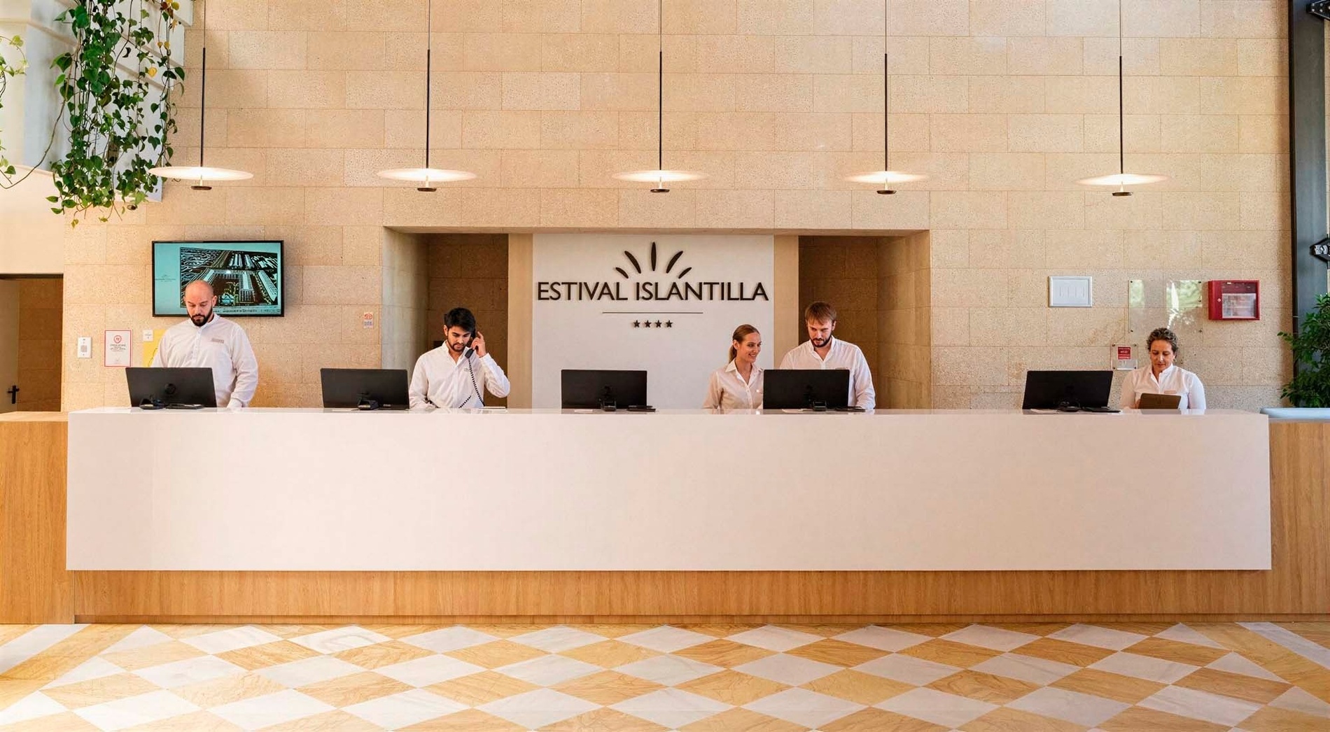 Five receptionists stand behind a modern white front desk in a hotel lobby with a large "Estival Islantilla" sign and light-colored stone walls.