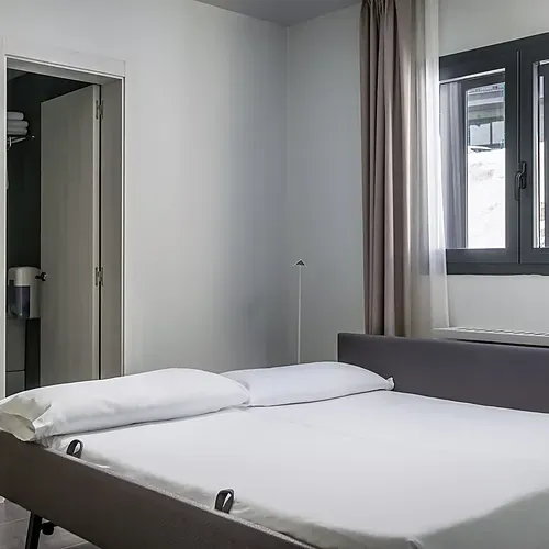 Comfortable hotel bed with white linens and a grey headboard, next to a closet and window. Hotel Cristina | Andorra | Estival Group | Official Web