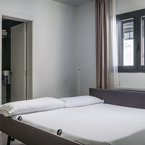 Comfortable hotel bed with white linens and a grey headboard, next to a closet and window. Hotel Cristina | Andorra | Estival Group | Official Web