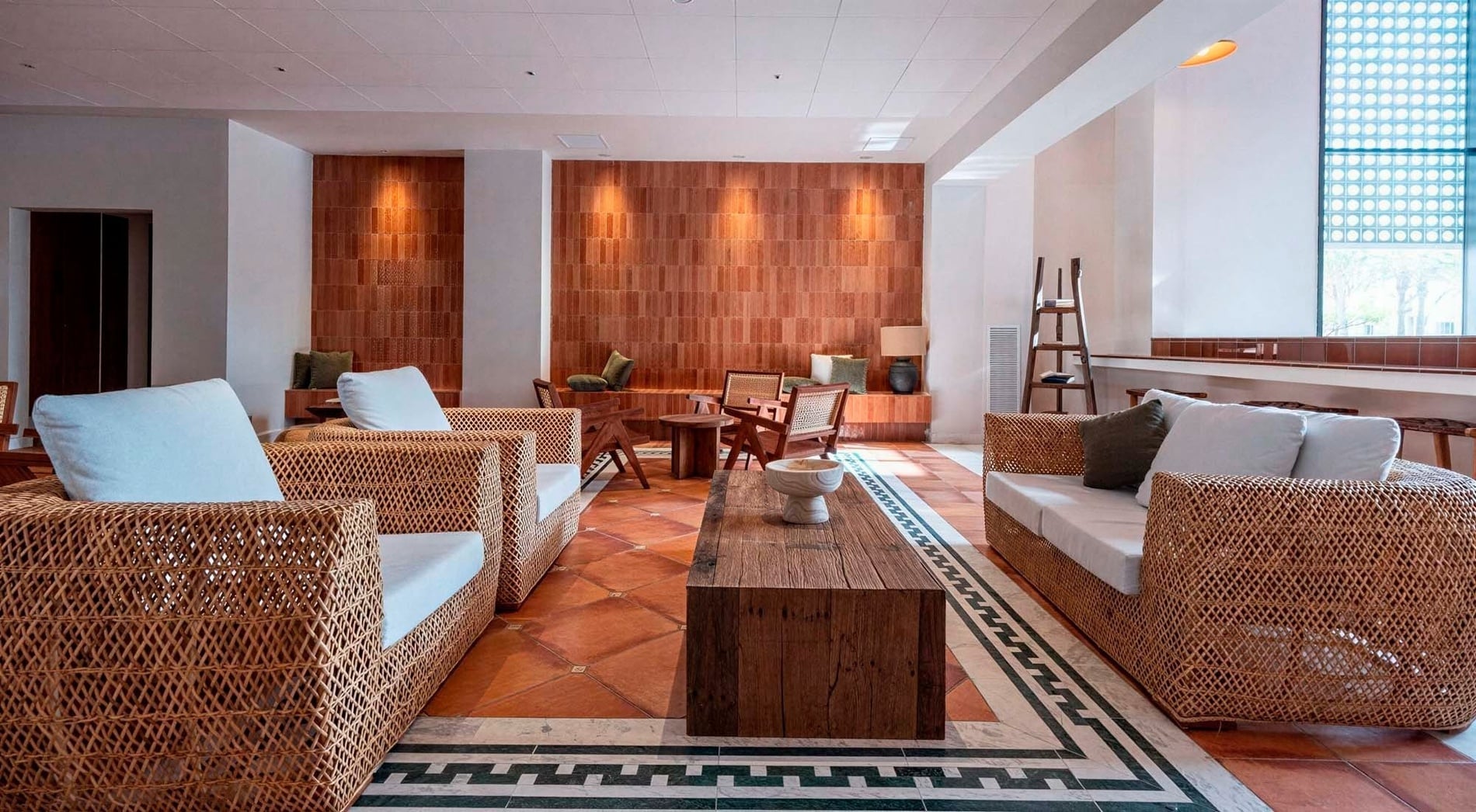 A cozy and naturally-lit lounge area features woven wicker furniture, terracotta floor tiles, and a prominent accent wall of reddish-brown bricks.