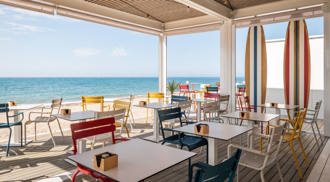 A vibrant beachfront cafe features colorful chairs and tables on a wooden deck, offering open views of the ocean and two decorative surfboards.