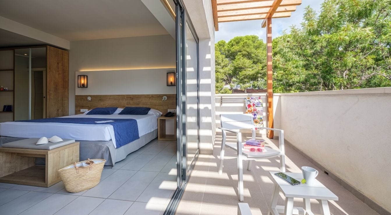 Moderna habitación de hotel con cama doble y balcón soleado amueblado con sillas blancas y vistas a la vegetación.