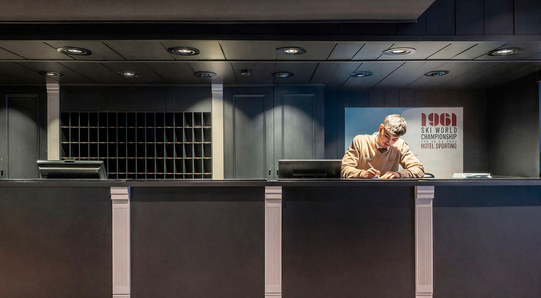 Un homme travaille à un comptoir de réception, avec des casiers et une affiche vintage de ski en arrière-plan.