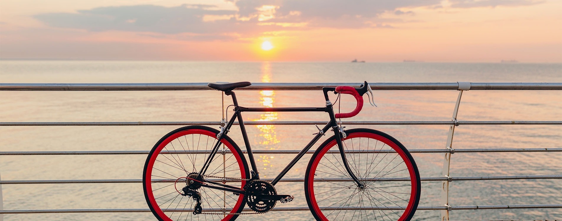 Una bicicleta negra amb rodes i manillar vermells està recolzada en una barana davant d'un mar tranquil amb el sol ponent a l'horitzó.