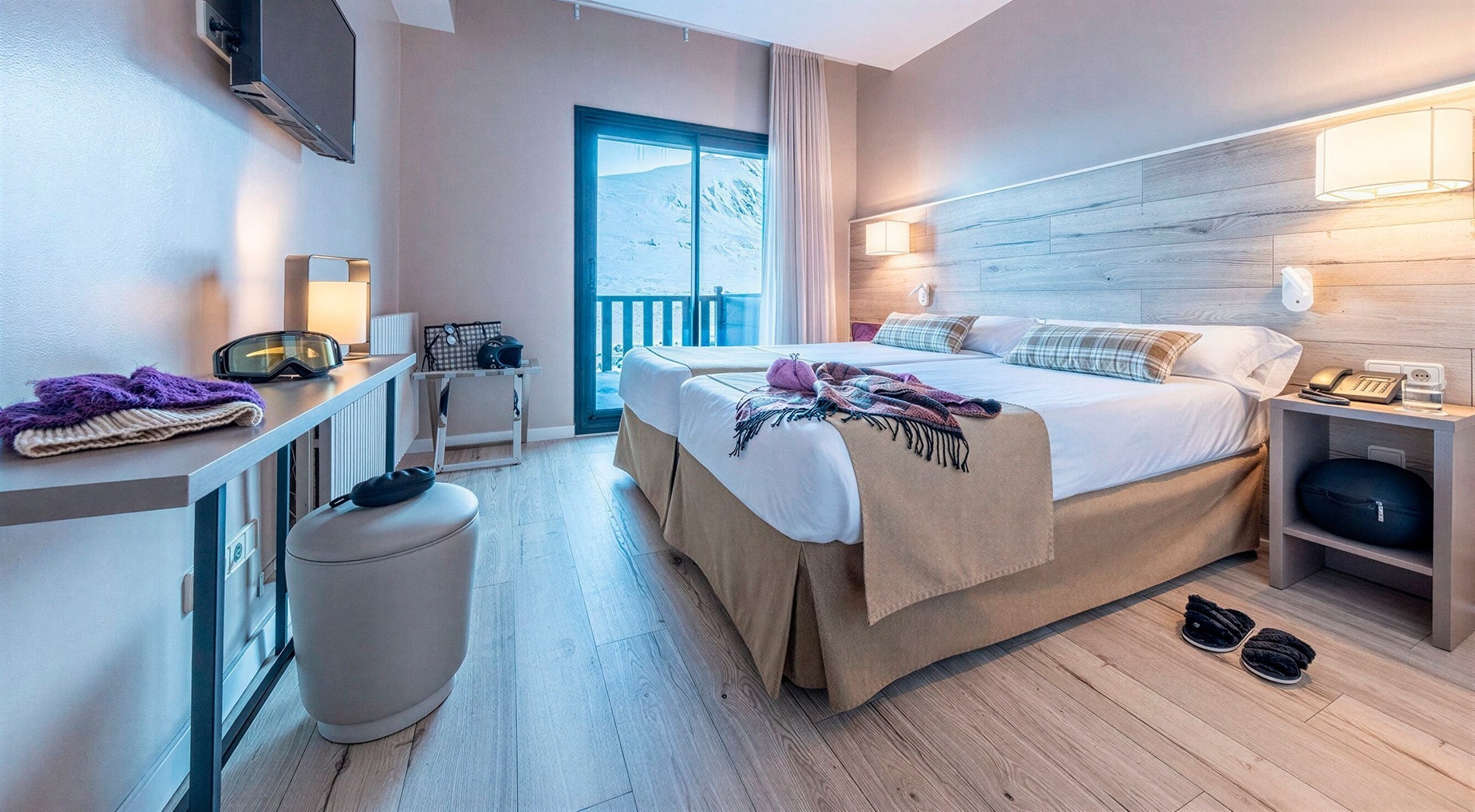 A modern and cozy hotel room, featuring two beds, ski gear, and a balcony with a view of snowy mountains, is illuminated by natural and artificial light.