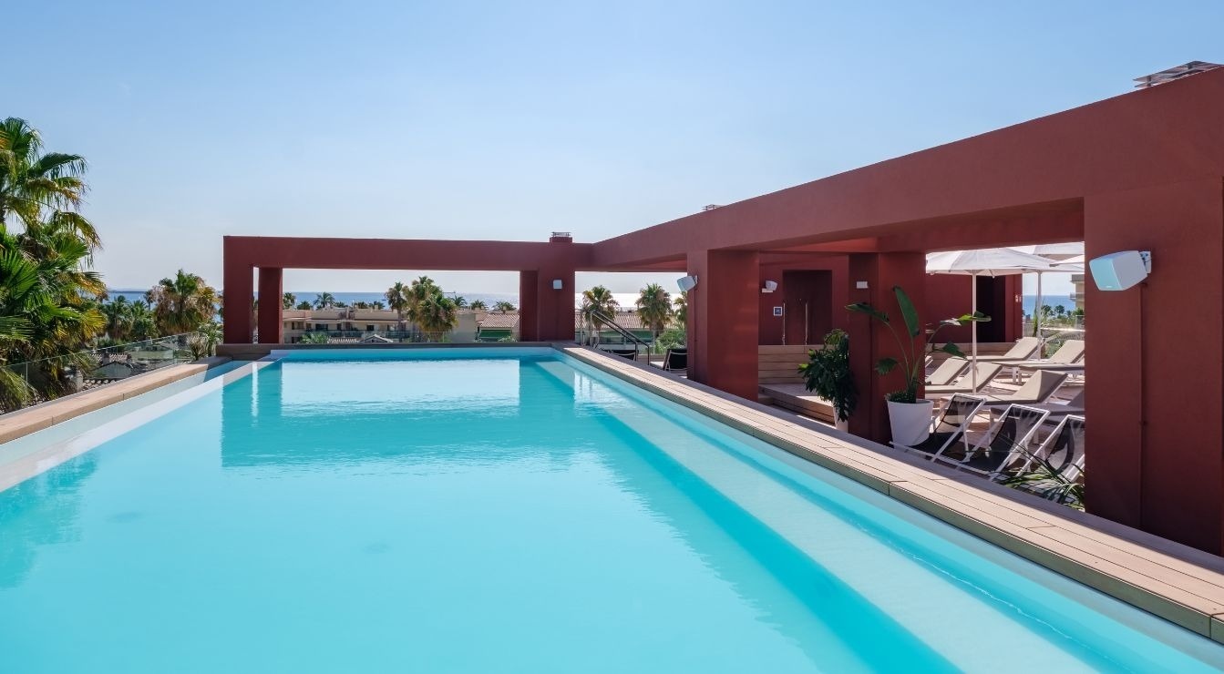 A bright blue rooftop swimming pool is framed by a terracotta-colored building, with palm trees and the sea visible in the background under a clear sky.