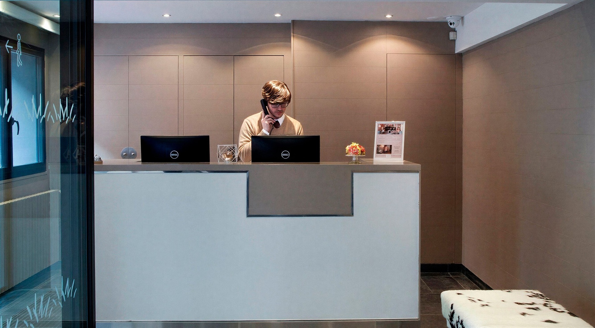 A man wearing glasses is speaking on a telephone behind a modern reception desk with two computer monitors and some decorative items in a neutral-toned room.