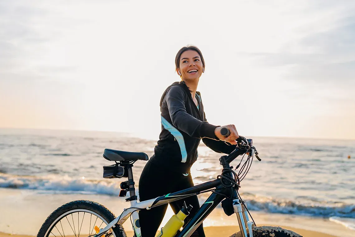 Mujer sonriente en bicicleta en la playa al amanecer, perfecta para ESTIVAL: Corporativa.