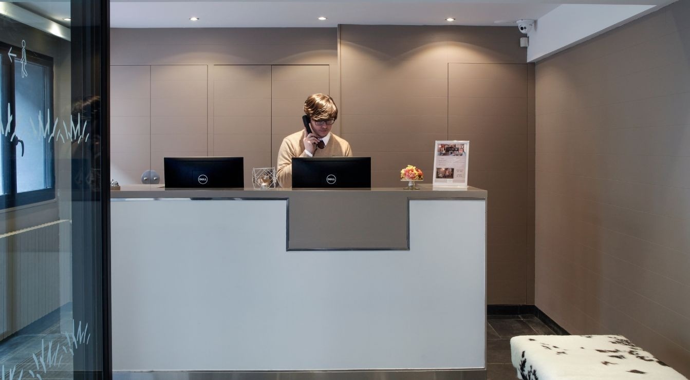 A man wearing glasses is speaking on a telephone behind a modern reception desk with two computer monitors and some decorative items in a neutral-toned room.