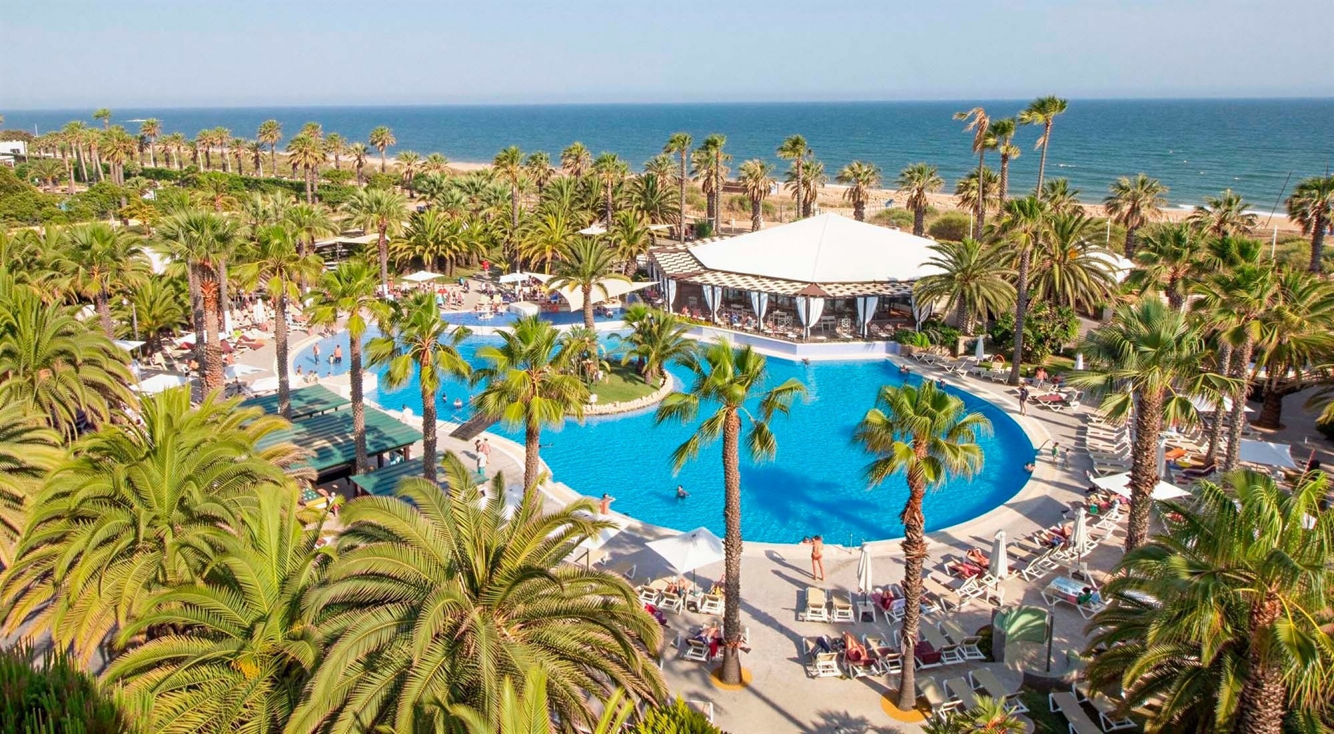 An aerial view captures a vibrant resort with a large, blue swimming pool surrounded by numerous palm trees, a white-canopied restaurant, and a sandy beach leading to the clear ocean under a sunny sky.