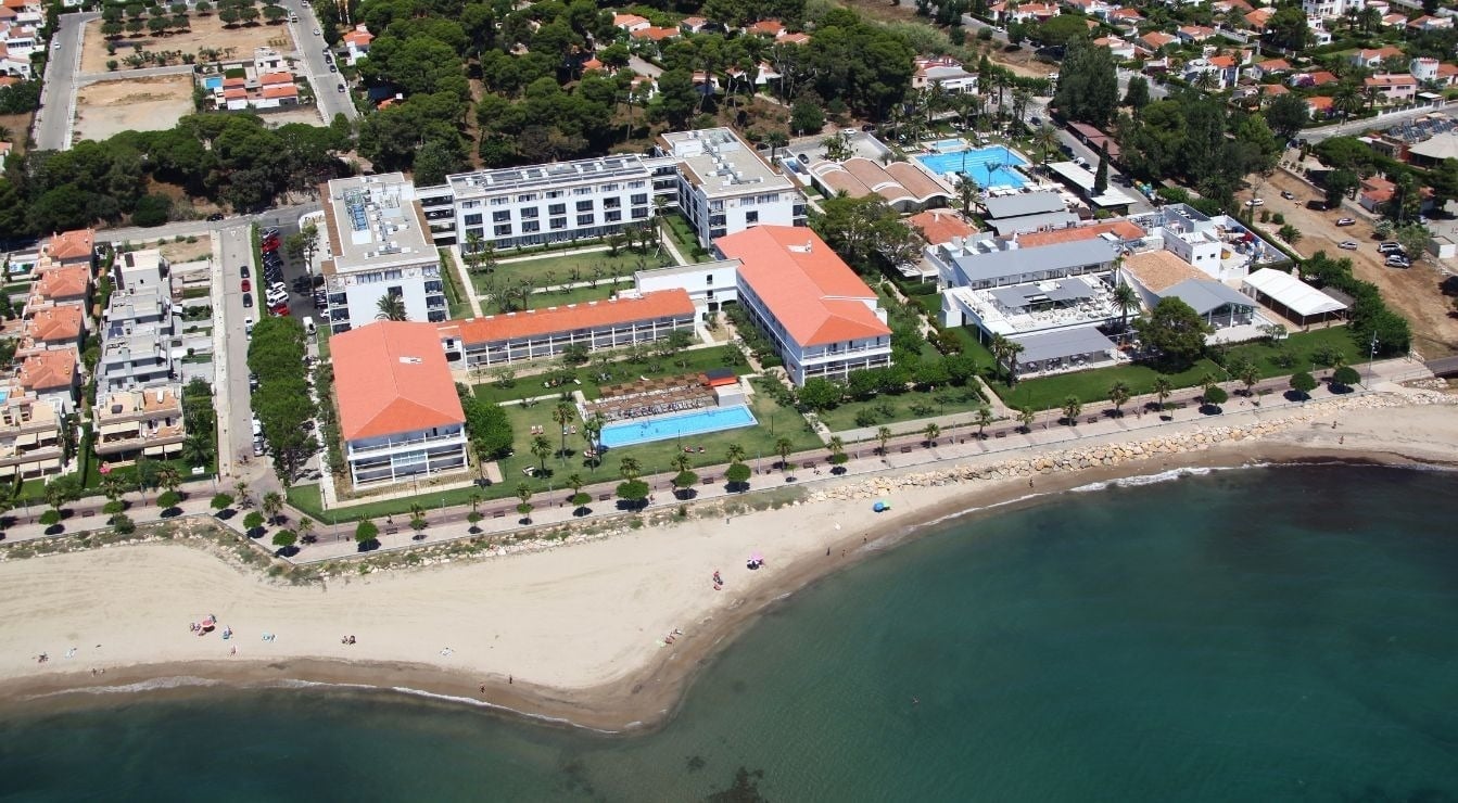 An aerial view captures a coastal resort complex featuring white buildings, orange roofs, multiple swimming pools, green lawns, a sandy beach, and clear blue sea.