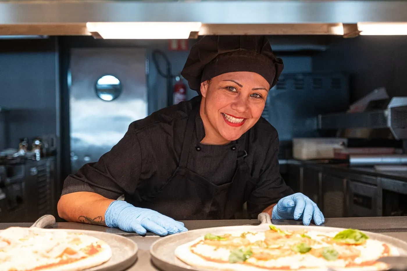 Smiling chef in ESTIVAL: Corporativa kitchen, preparing pizzas with gloves.