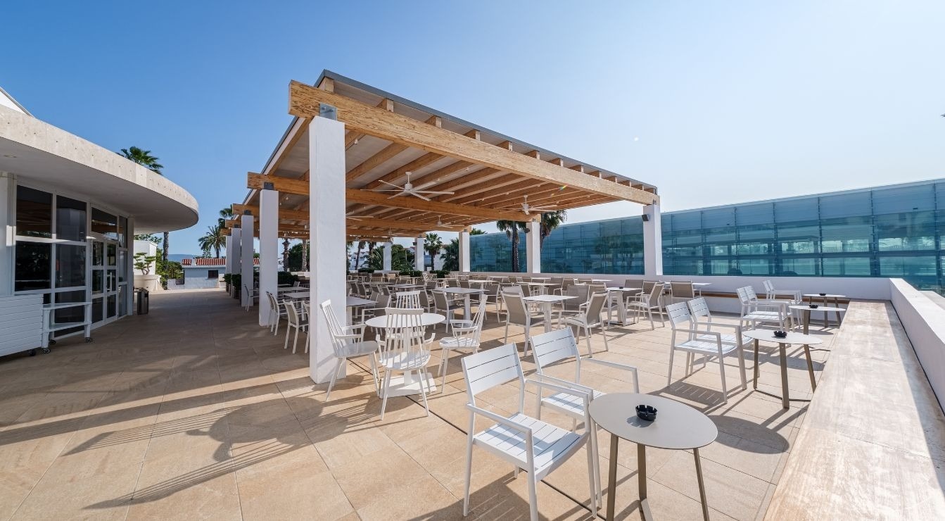 A sunny outdoor terrace features numerous white tables and chairs beneath a wooden pergola, casting long shadows, with contemporary buildings and palm trees visible in the background under a clear blue sky.