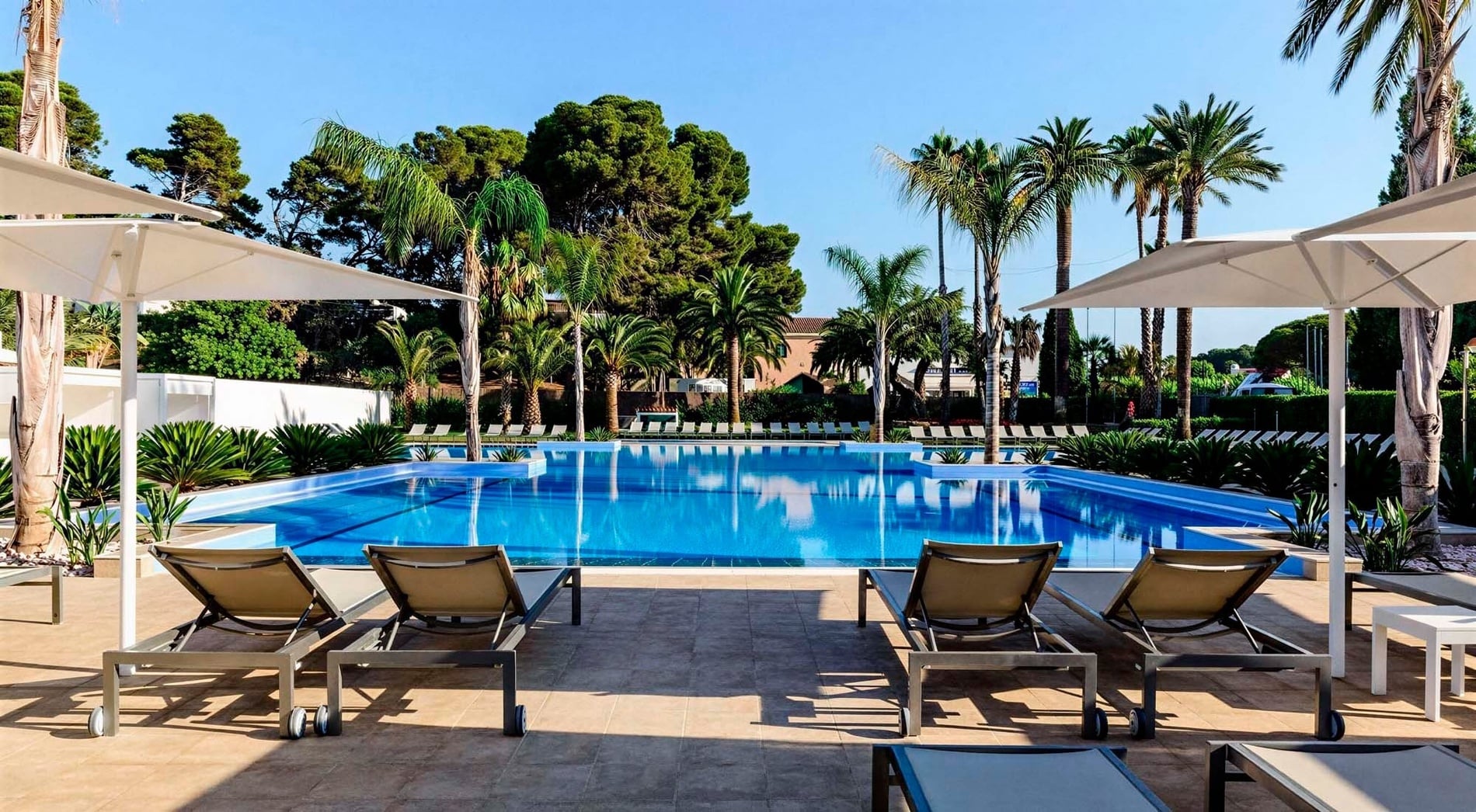 Una piscina azul brillante con tumbonas y sombrillas blancas, rodeada de palmeras y abundante vegetación bajo un cielo azul claro.