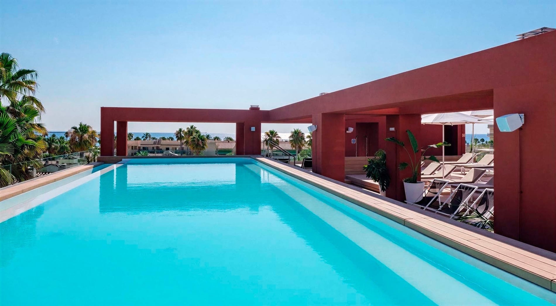 A bright blue rooftop swimming pool is framed by a terracotta-colored building, with palm trees and the sea visible in the background under a clear sky.
