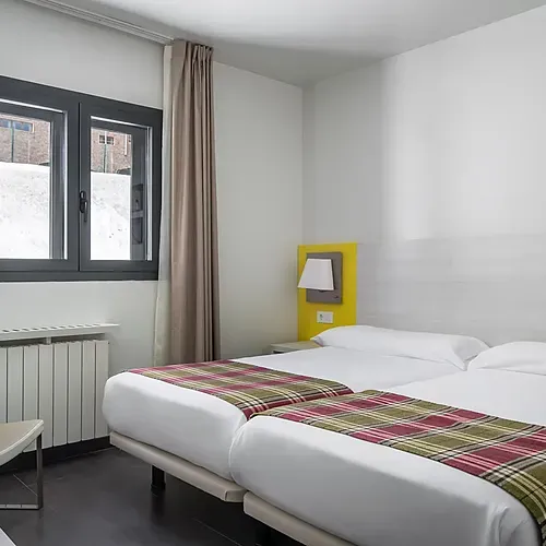 Modern hotel room with two beds, tartan throws, window view, and cozy seating. Hotel Cristina | Andorra | Estival Group | Official Web