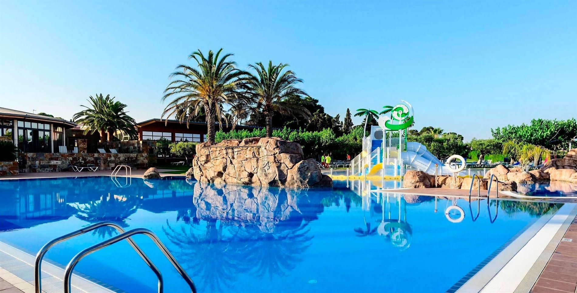 Una piscina blava radiant amb palmeres i edificis al fons, complementada amb una formació rocosa i una zona de jocs aquàtics per a nens, sota un cel clar.
