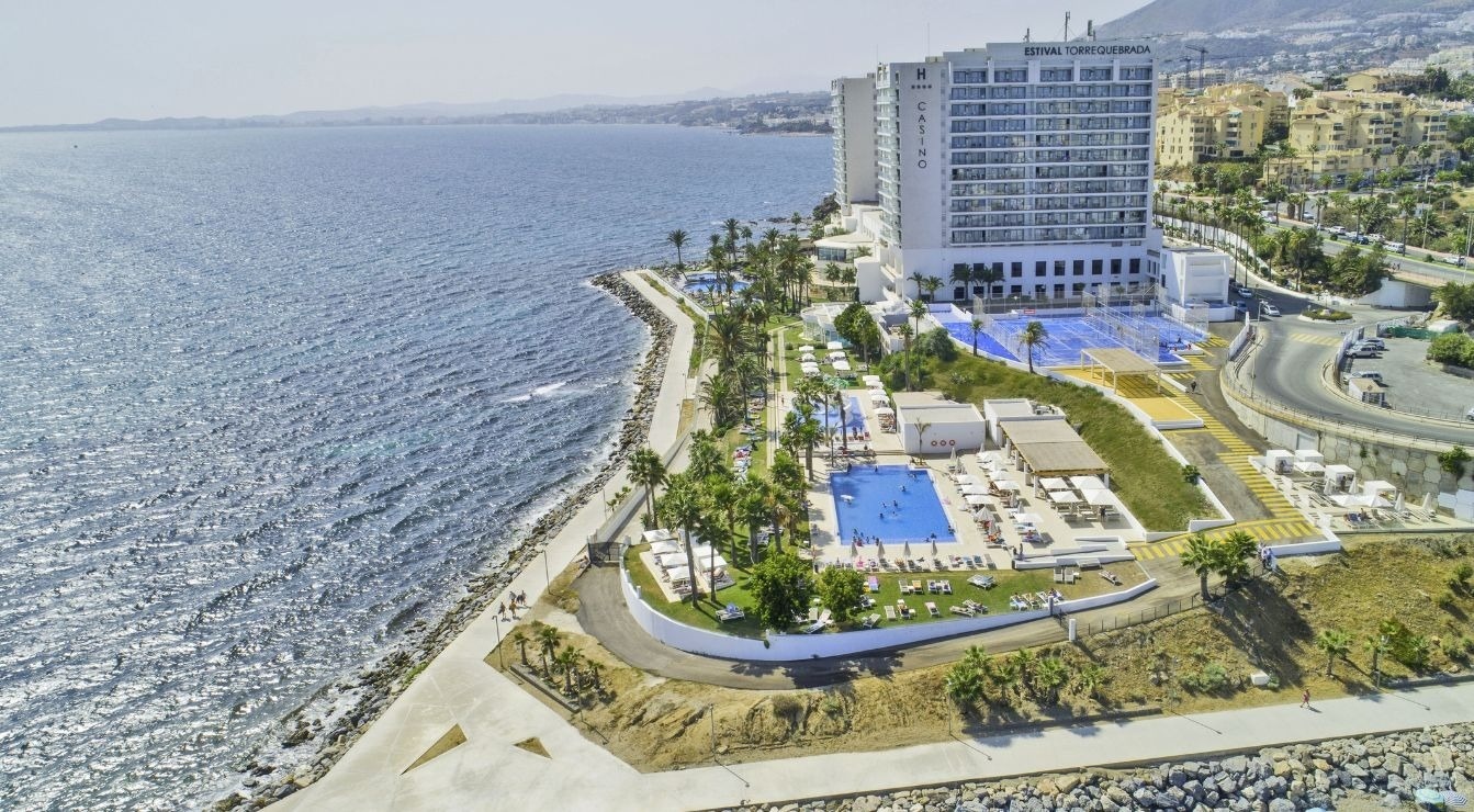 An aerial view captures a large white resort complex with swimming pools and sports courts situated along a rocky coastline next to a sparkling blue sea, with a road and other buildings ascending a hill in the background.