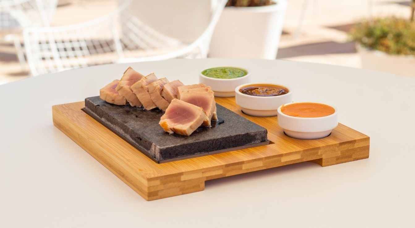 Des tranches de thon mi-cuit sont servies sur une plaque de pierre avec trois coupelles de sauces, le tout présenté sur un plateau en bambou sur une table blanche.