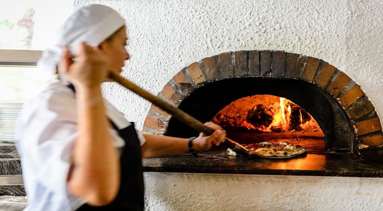 Un cocinero introduce una pizza en un horno de leña tradicional con fuego visible usando una pala.