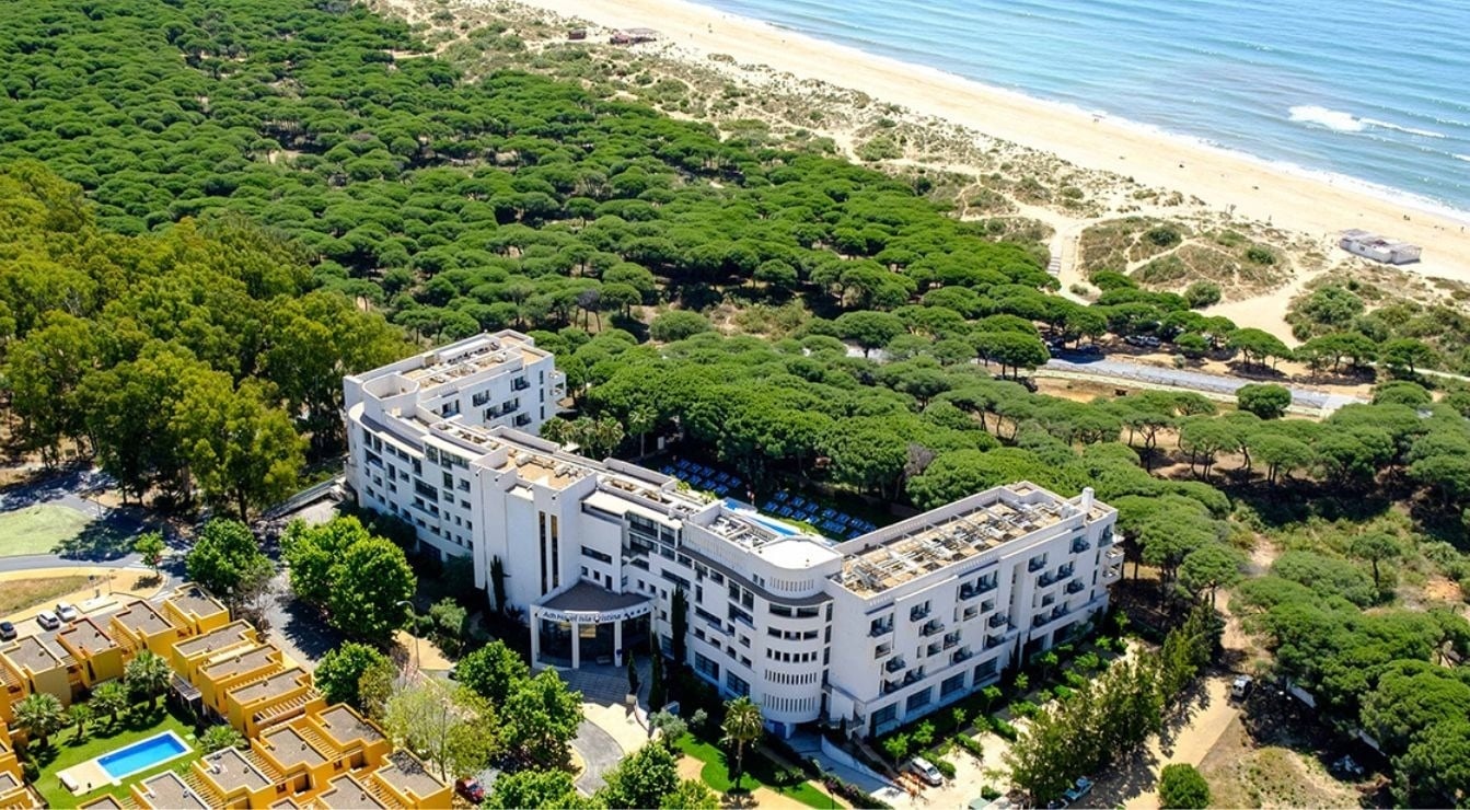 Une vue aérienne présente un complexe hôtelier moderne et des logements, entourés d'une forêt luxuriante, bordant une plage de sable fin et l'océan.