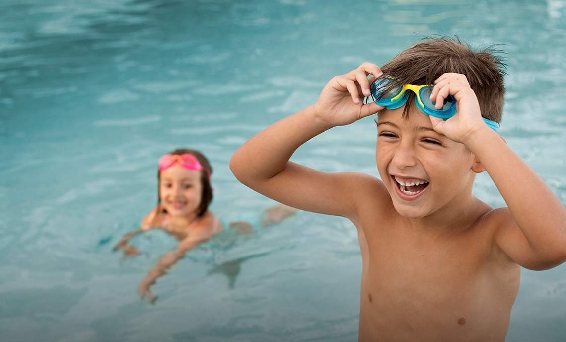 a boy wearing goggles smiles while a girl swims in the background