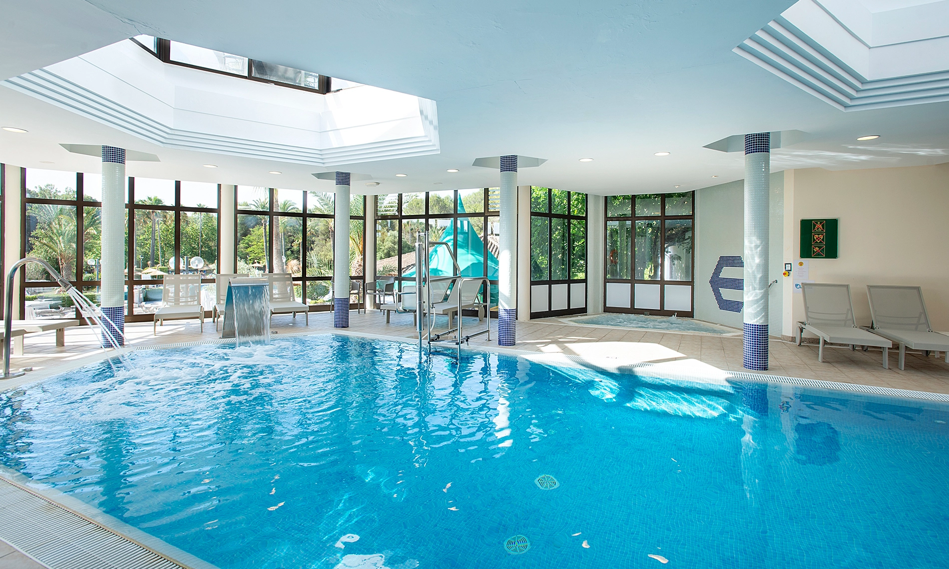 Piscina interior lluminosa de Hotel Exagon Park amb tumbones i cascades, vista al jardí exterior.