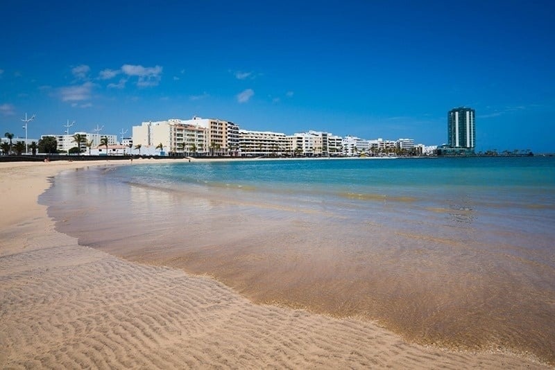 Una hermosa playa de arena clara y mar azul se extiende frente a una ciudad con edificios costeros y un rascacielos prominente, todo bajo un cielo azul despejado.