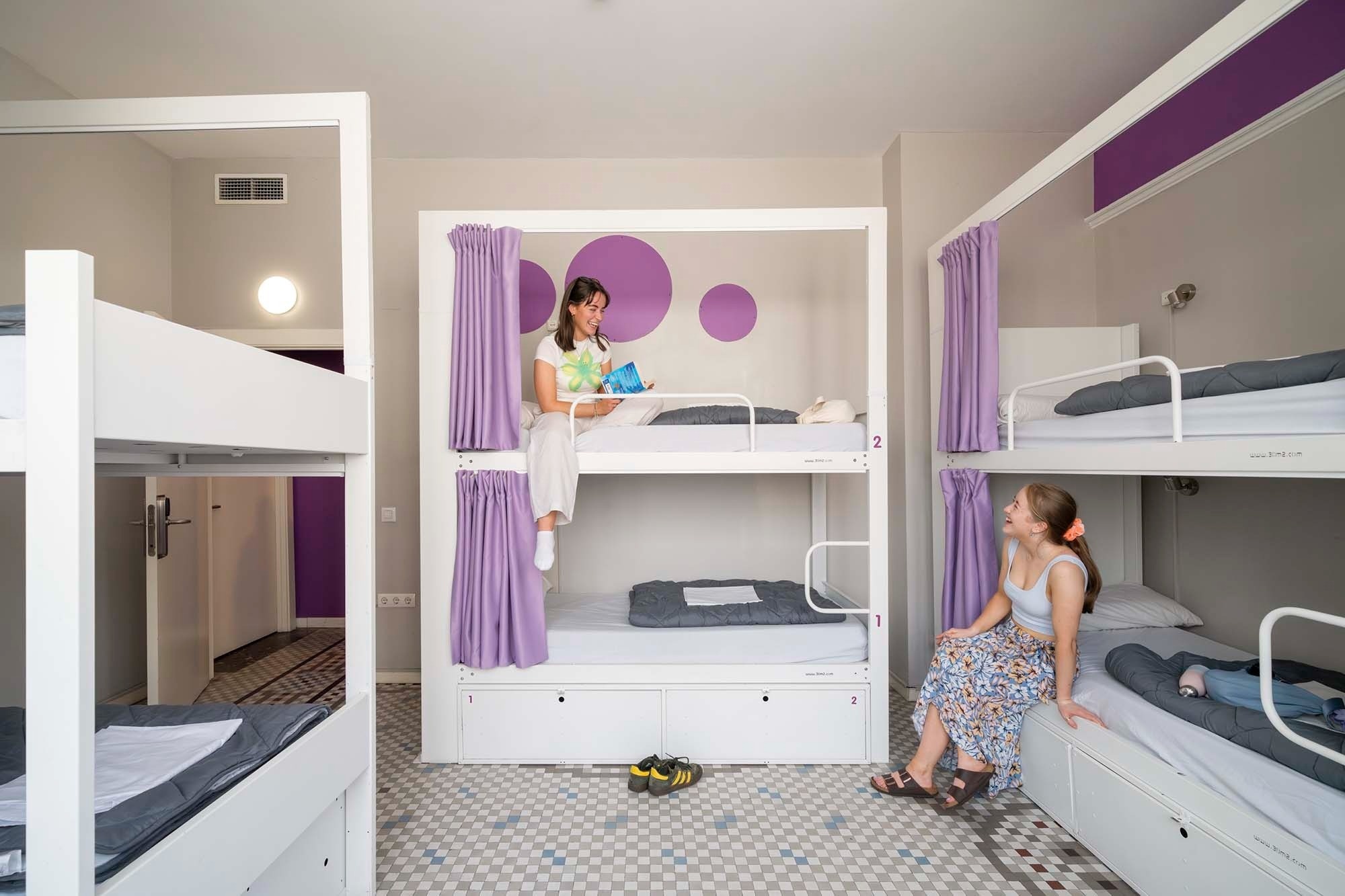 Bunk beds with purple curtains and relaxed girls. Feet up! Modern hostel vibe.