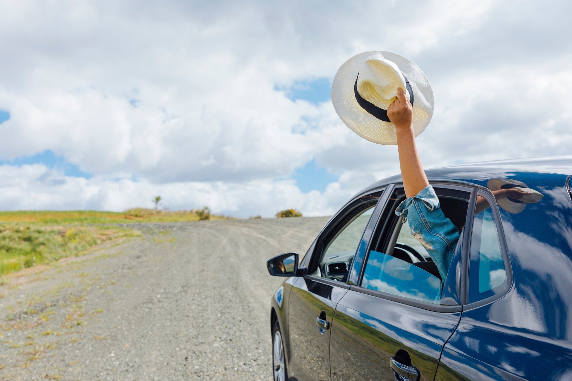 Un brazo con un sombrero asoma por la ventanilla de un coche oscuro en un camino de tierra bajo un cielo nublado.