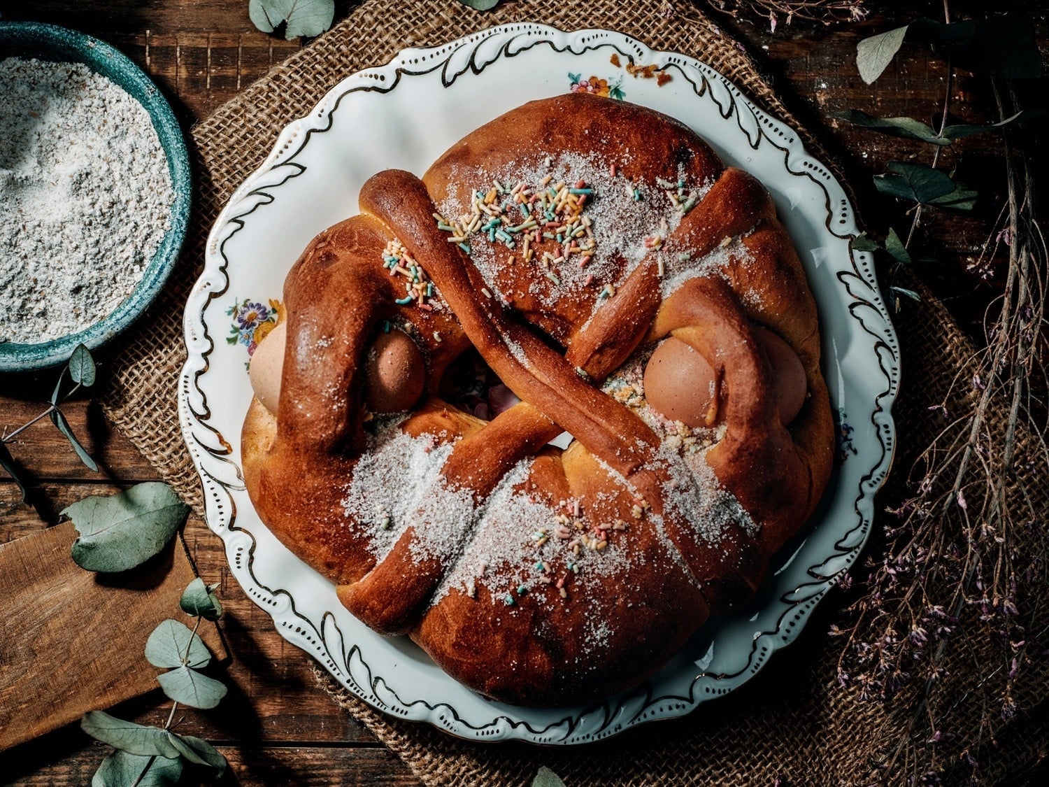A festive, braided Easter bread (likely a folar) embedded with whole eggs, dusted with sugar and colorful sprinkles, is beautifully presented on a decorative plate on a rustic wooden table with burlap, a bowl of flour, and dried foliage.