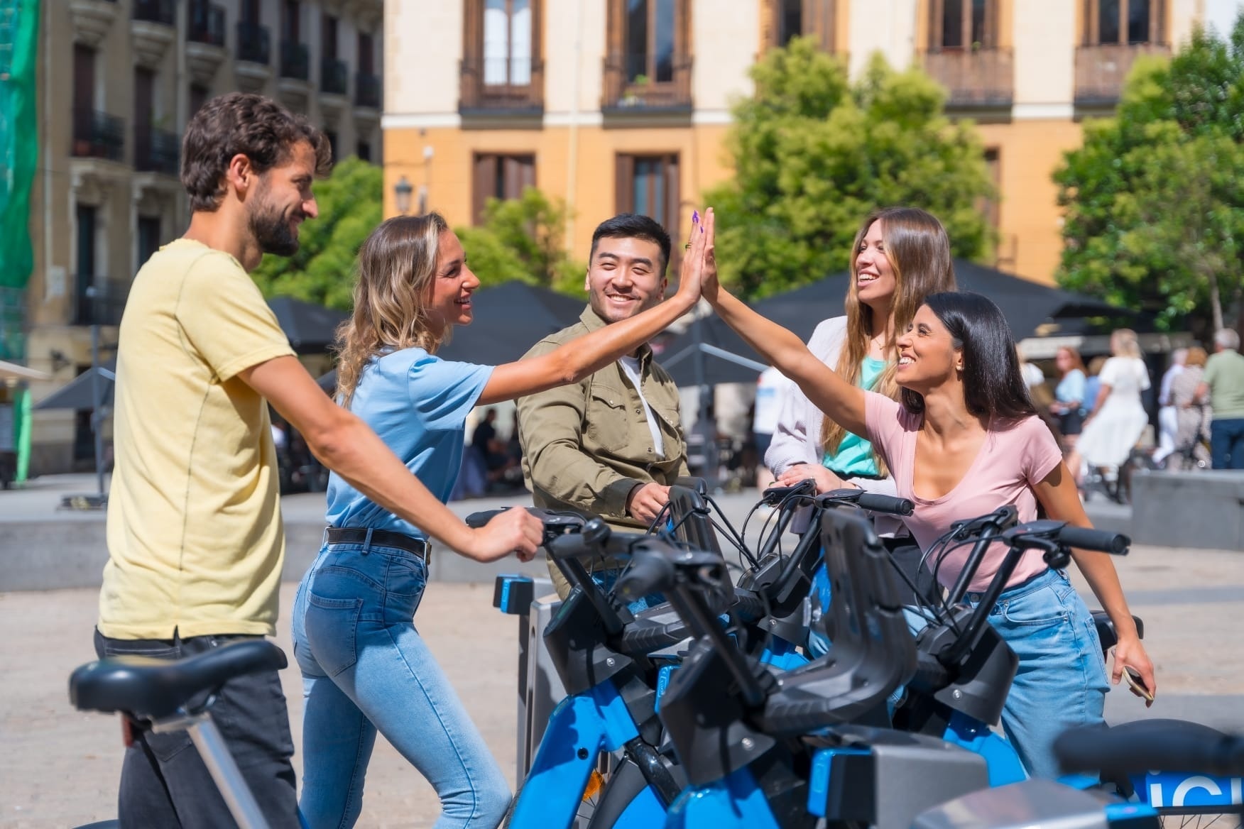 Happy friends high-fiving at a city bike rental station, enjoying urban adventure.