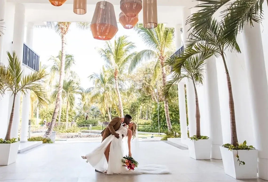 Newlyweds kissing at The Fives Corpo, a tropical destination with palm trees and hanging lights.