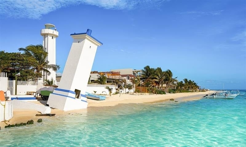 This vibrant image shows a standing lighthouse alongside a heavily leaning lighthouse on a tropical sandy beach with clear turquoise waters, palm trees, and coastal homes under a bright blue sky. Puerto Morelos, Riviera Maya