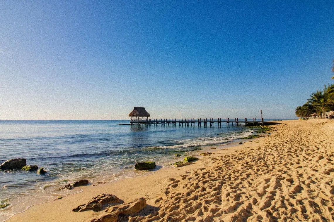 Tropical The Fives Corpo: pier with thatched-roof cabana over calm sea, sandy beach, palm trees.