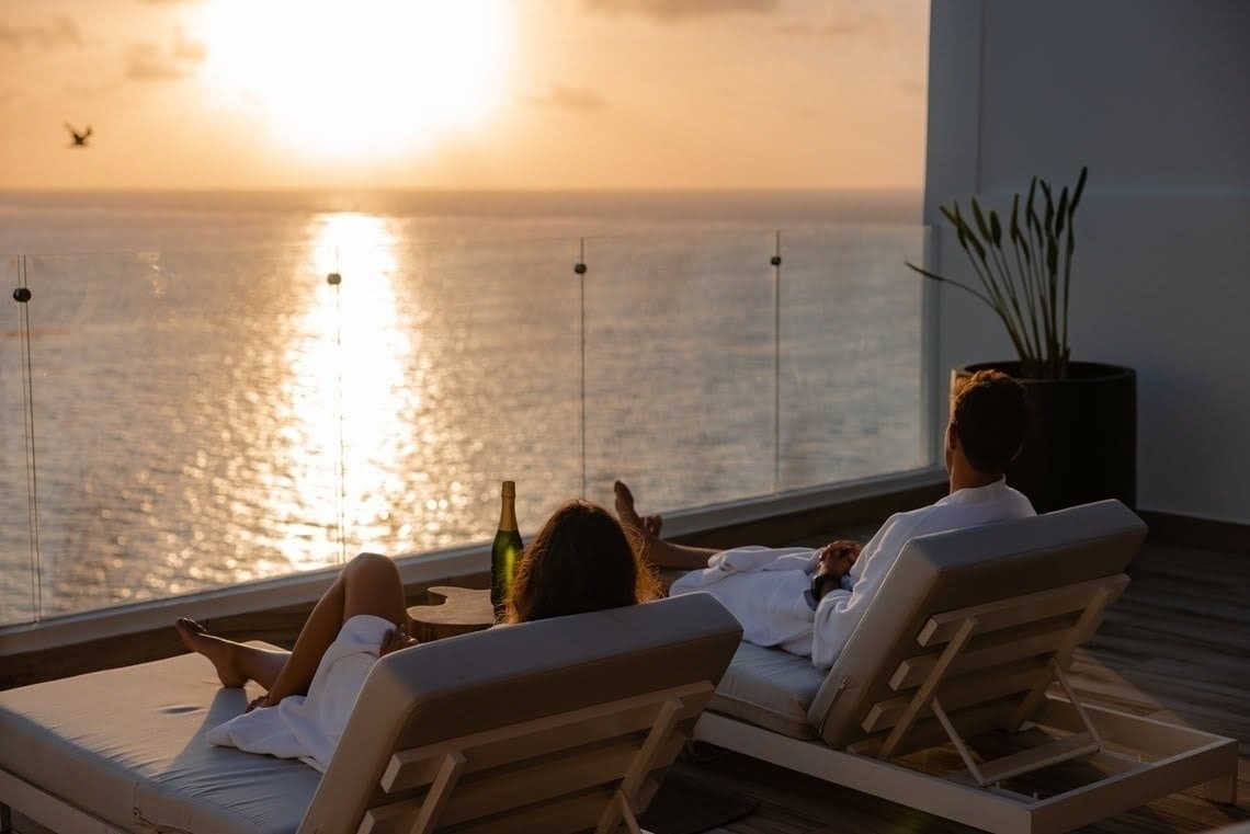 Luxury hotel balcony with couple in robes enjoying a romantic ocean sunset view and champagne.