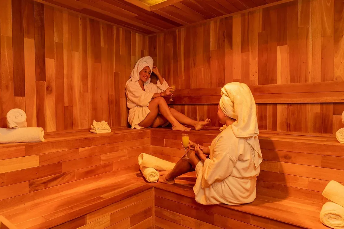 Two women enjoy drinks in The Fives Corpo sauna, featuring towels and robes.