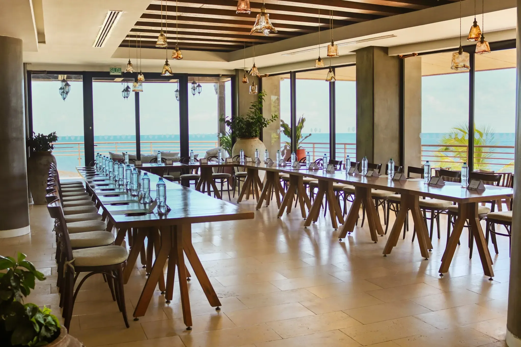 Salón de conferencias con vista al mar, mesas largas dispuestas, sillas de madera y lámparas colgantes. Perfecto para tu The Fives Corpo.