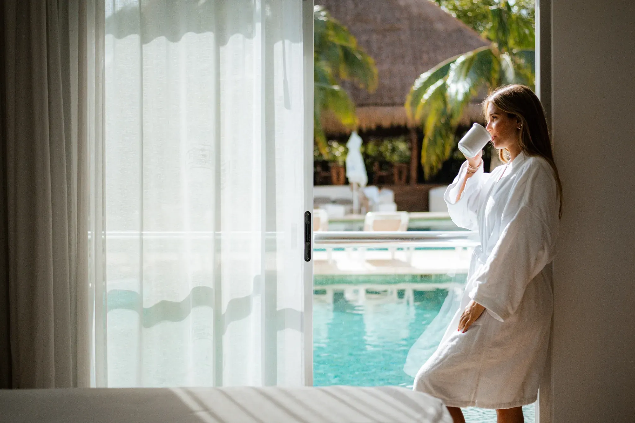 Woman in white robe drinks coffee by pool, enjoying The Fives Corpo.