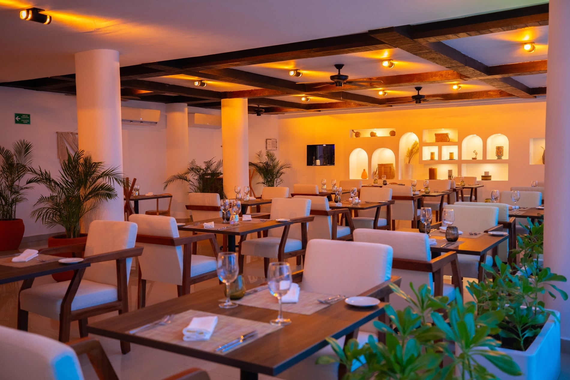 Modern hotel dining room with warm lighting, wood accents, set tables, and lush green plants.